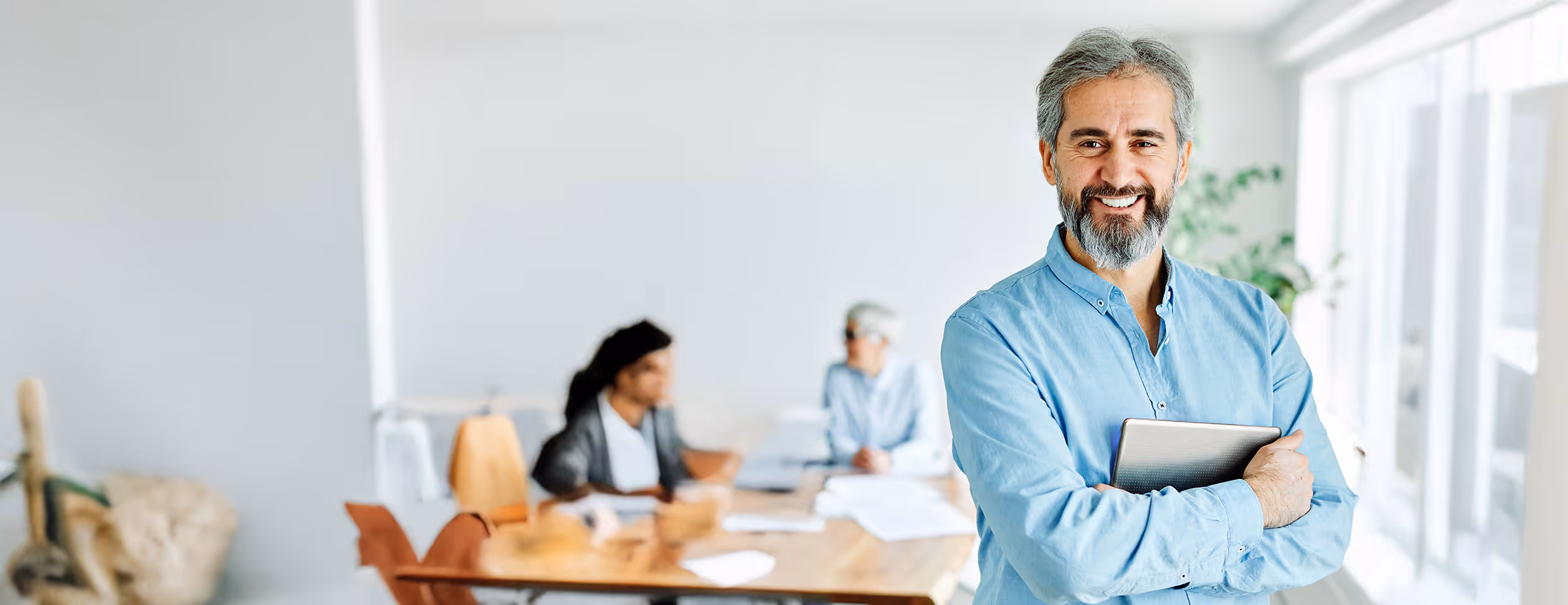 Lächelnder Mann mit grauem Bart und blauem Hemd hält Tablett, im Hintergrund zwei unscharfe Personen an einem Tisch.