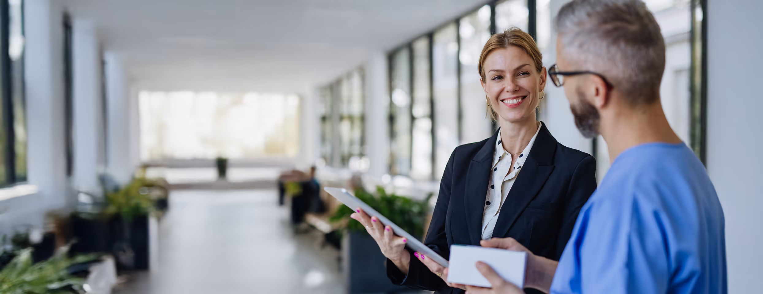 Frau in schwarzem Blazer lächelt einen Mann in blauem medizinischem Kittel an, während sie in einem hellen Flur ein Tablet hält.
