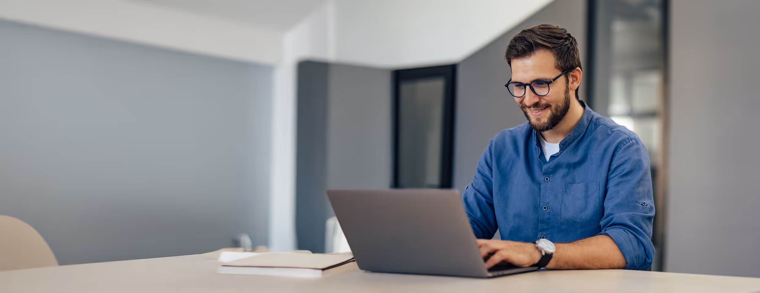 Junger Mann mit Brille und blauem Hemd arbeitet lächelnd an einem Laptop an einem weißen Tisch in einem modernen Büro.