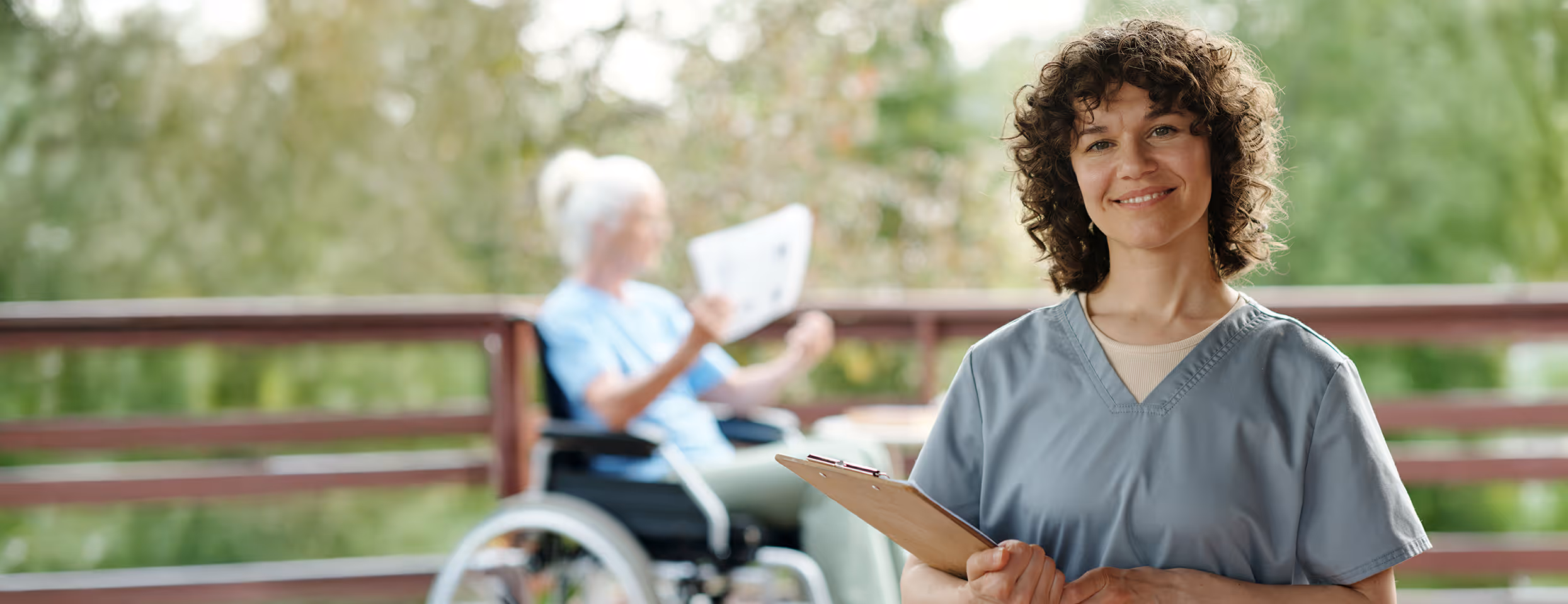 Pflegekraft mit Klemmbrett lächelnd vor älterer Frau im Rollstuhl auf einer Terrasse.