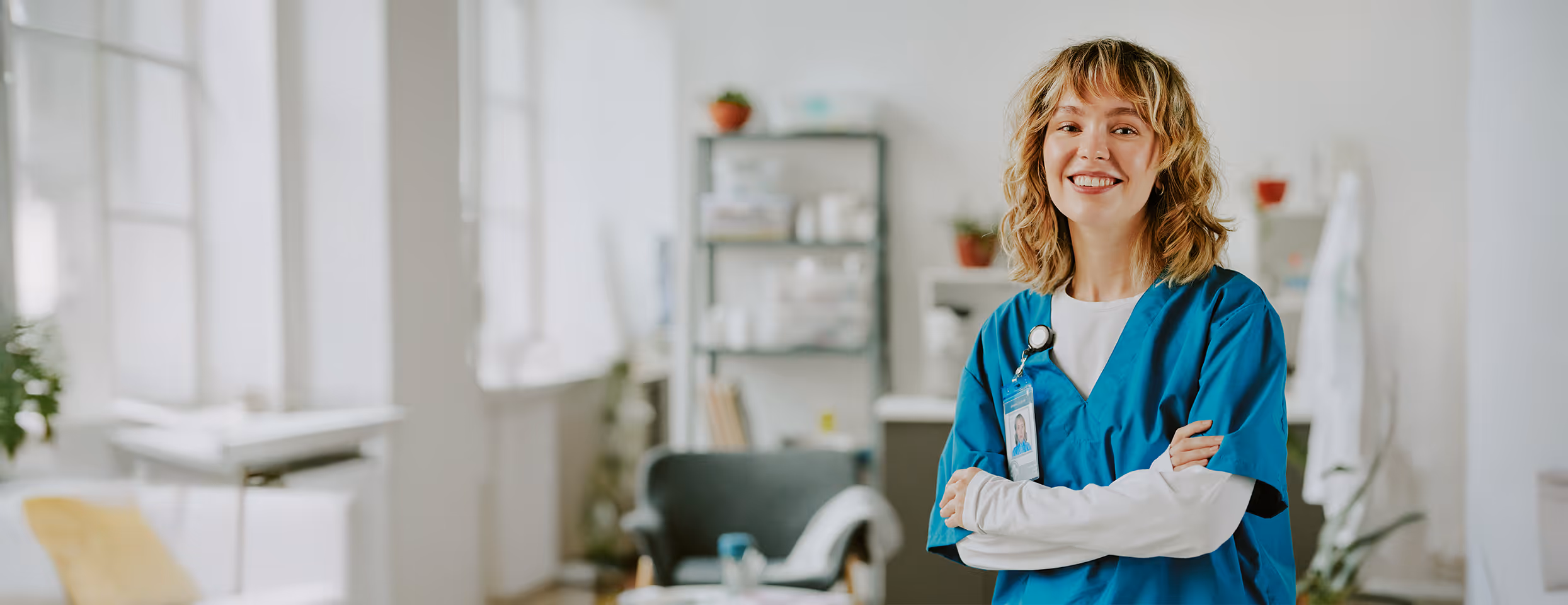 Lächelnde Krankenschwester in blauer Arbeitskleidung mit verschränkten Armen in einem hellen und modernen Behandlungsraum.