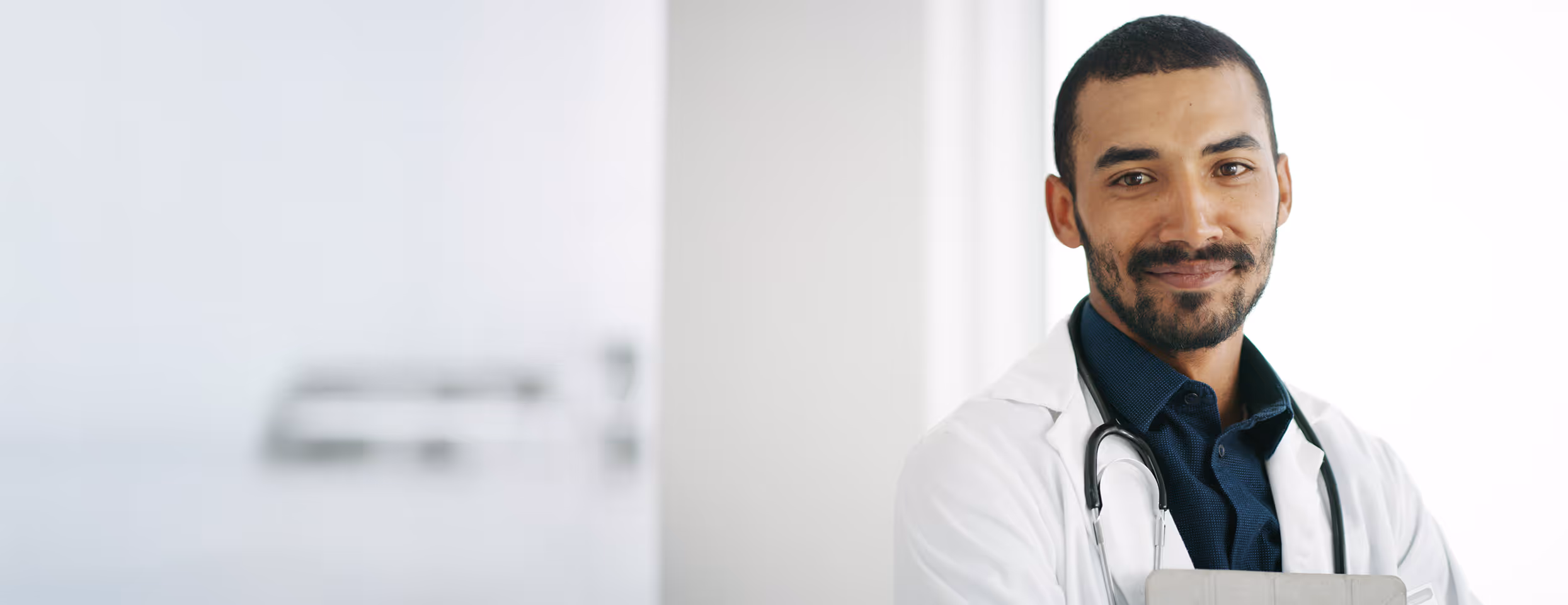 Lächelnder Arzt mit weißem Kittel und Stethoskop vor hellem, unscharfem Hintergrund.