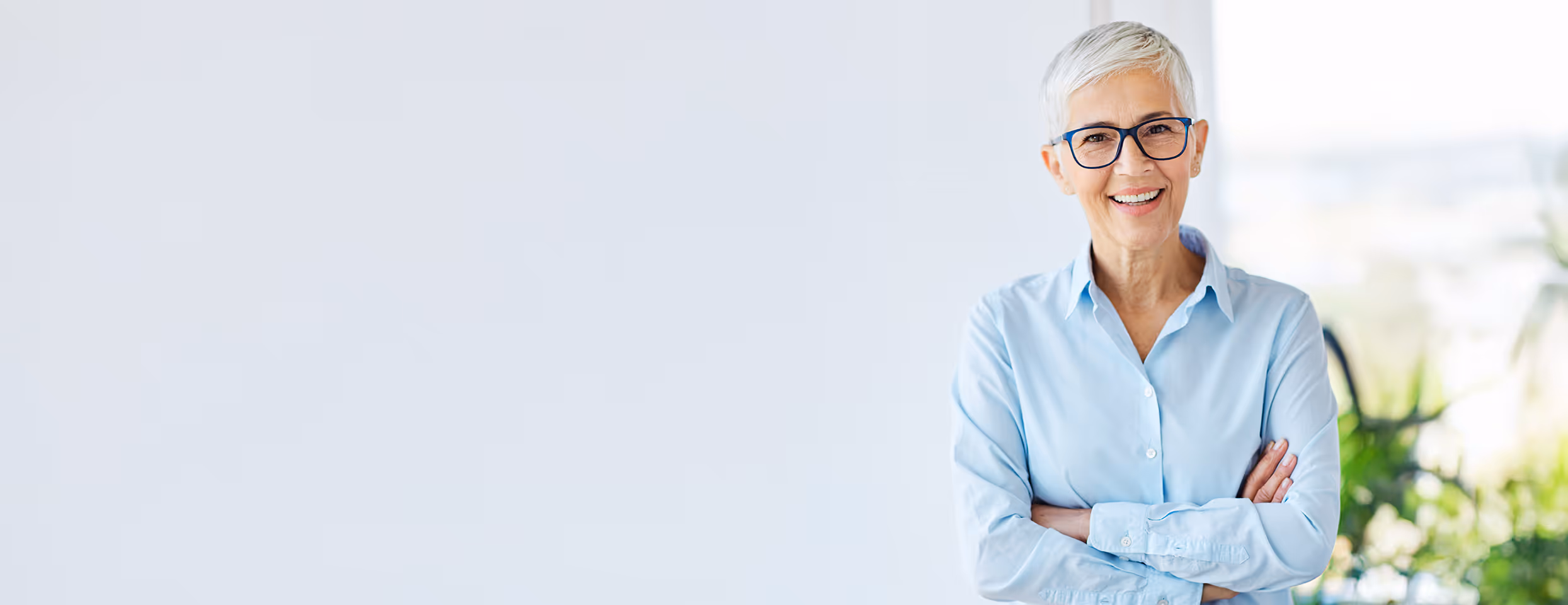 Ältere Frau mit kurzen weißen Haaren und blauer Brille lächelt und steht mit verschränkten Armen vor einem hellen Hintergrund.