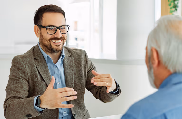Lächelnder Mann mit Brille erklärt etwas mit den Händen bei einem Gespräch mit älterem Mann.