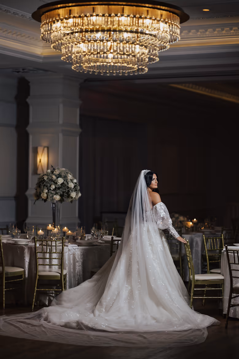 Bride in a long white gown and veil standing next to a chair under a grand chandelier in a decorated room.