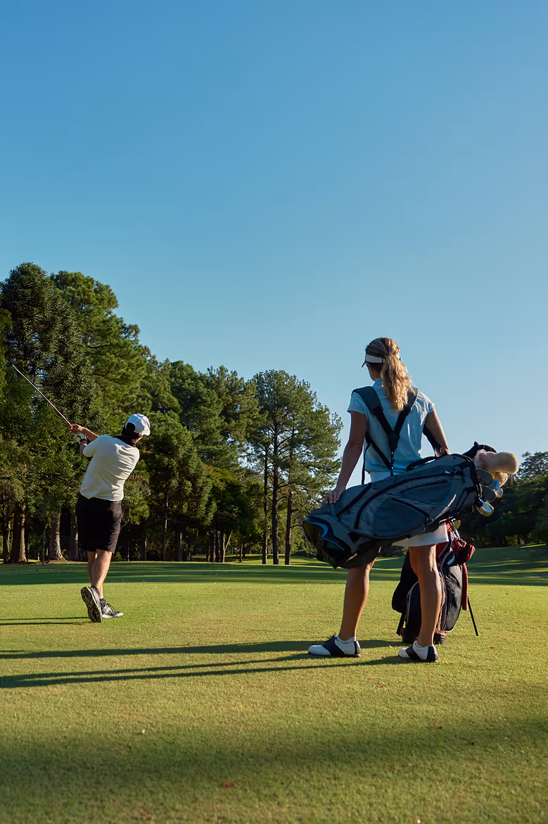 Man swings golf club while woman with golf bag watches on lush green golf course.