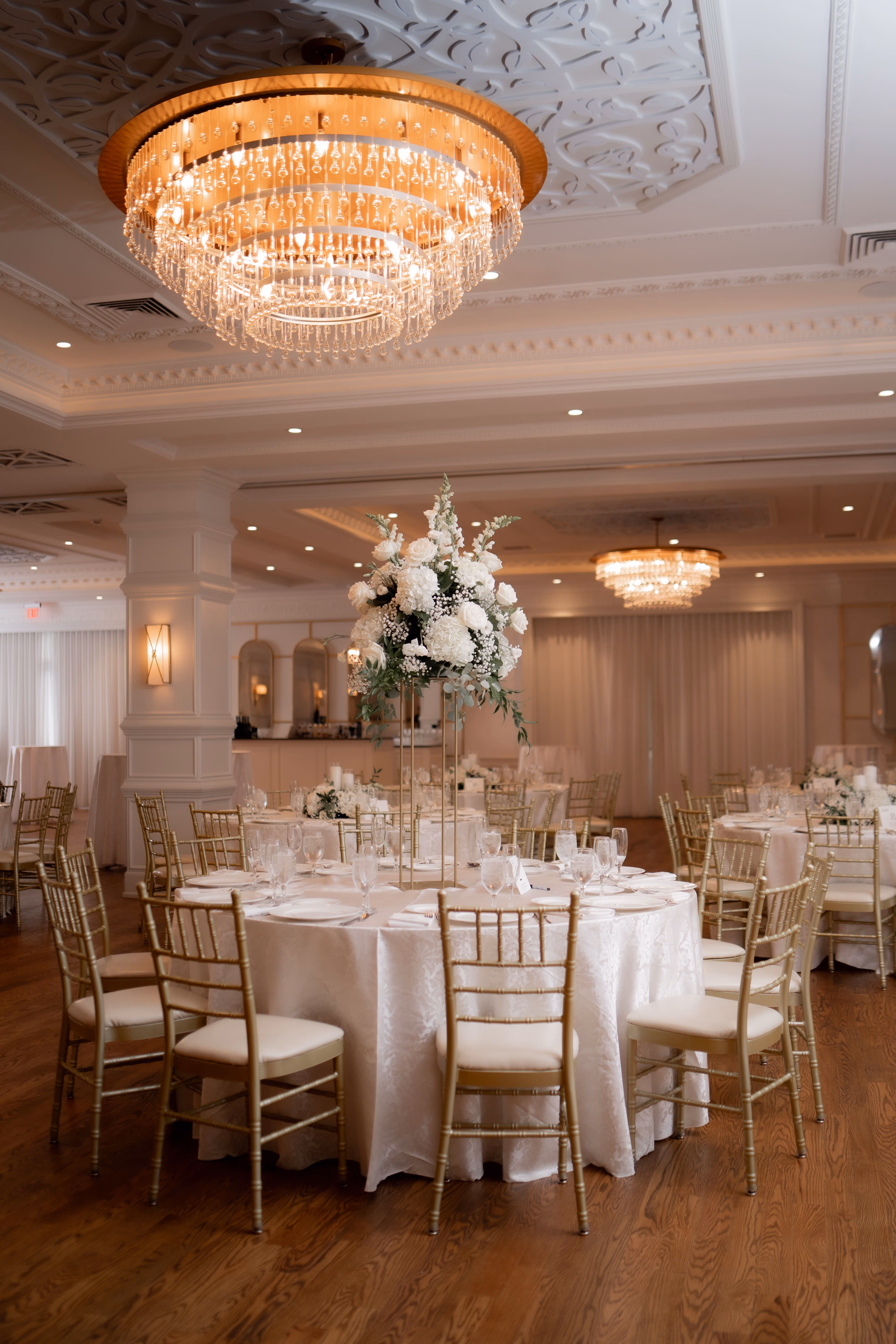 Elegant banquet room with round tables, gold chairs, white floral centerpieces, and crystal chandeliers.