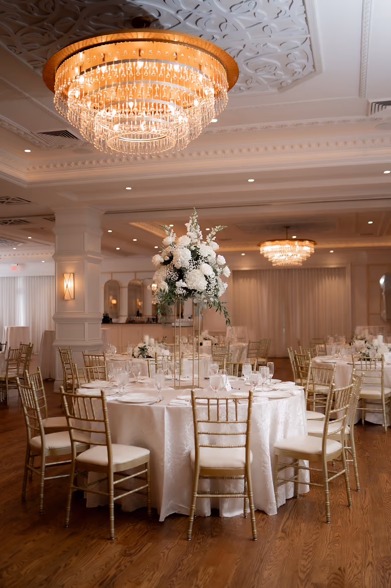 Elegant round table with white floral centerpiece and gold chairs in a banquet hall with chandeliers.