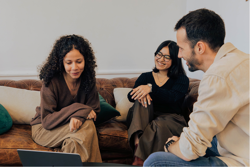 Three people sitting on a brown leather sofa, looking at a laptop and smiling.