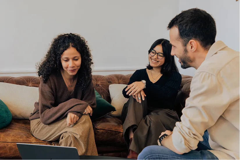 Three people sitting on a brown leather sofa, looking at a laptop and smiling.