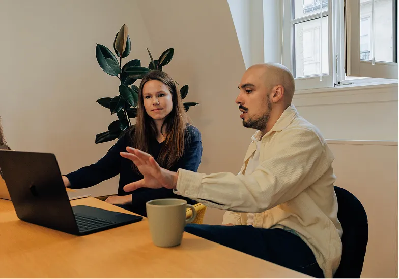 Man and woman sitting at a table, discussing while looking at a laptop in a bright room.