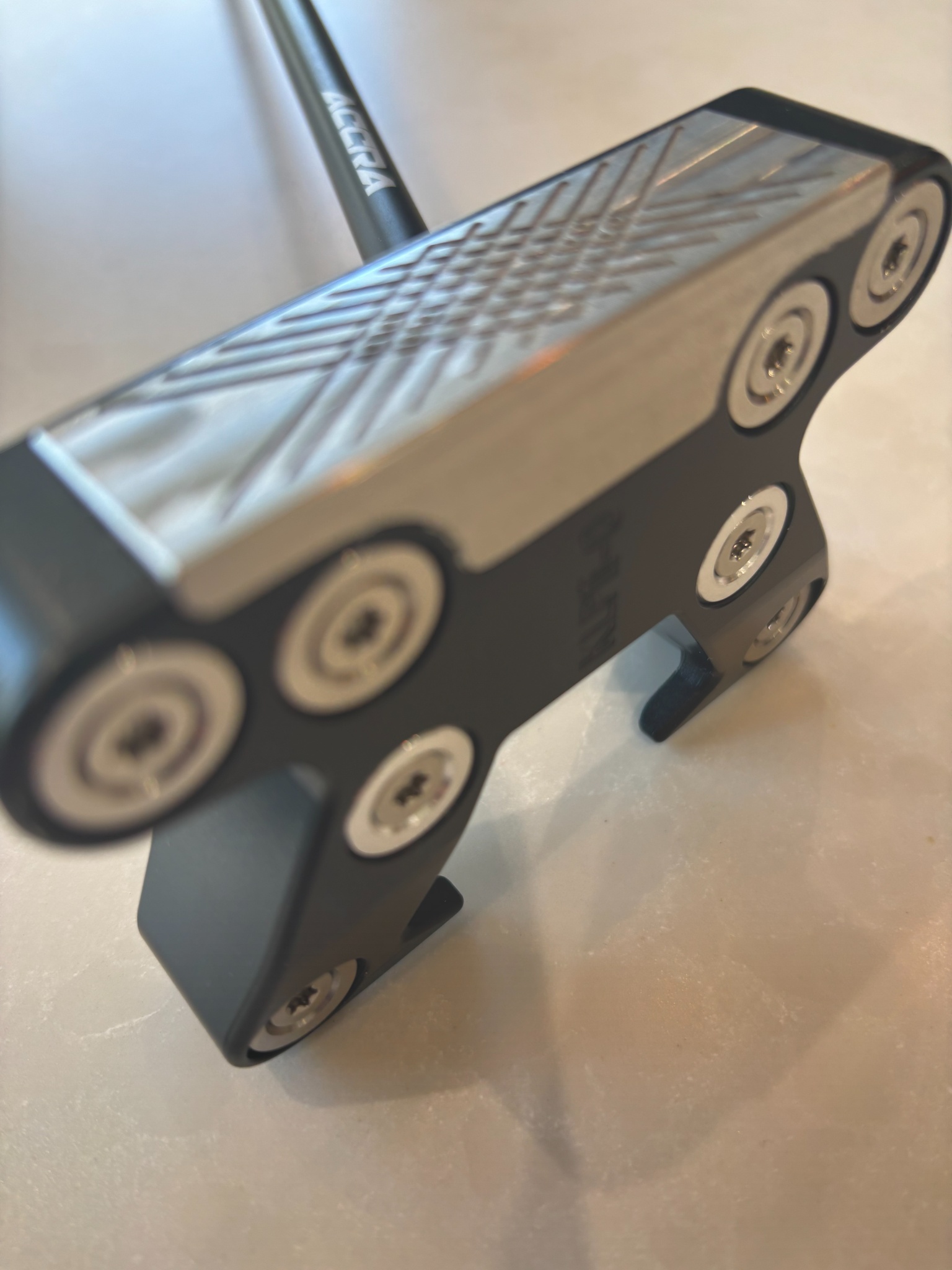 Close-up of a golf putter head with screws and patterned metal face on a light surface.