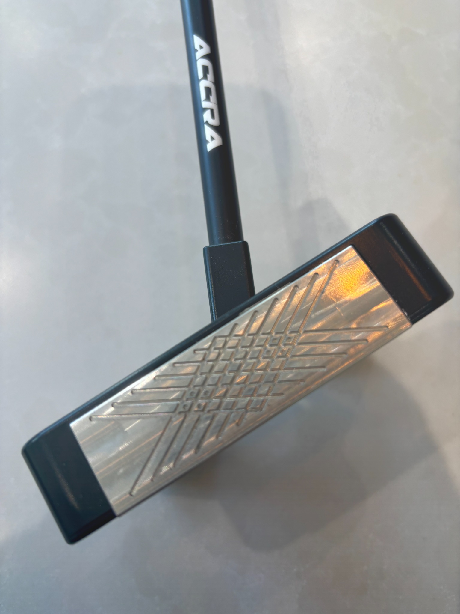 Close-up of a golf putter head with a geometric pattern and a black shaft marked Accra.