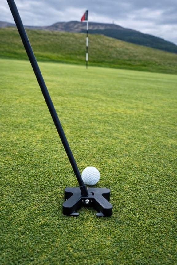 Golf ball positioned by a putter on a green with a flag and hills in the background.