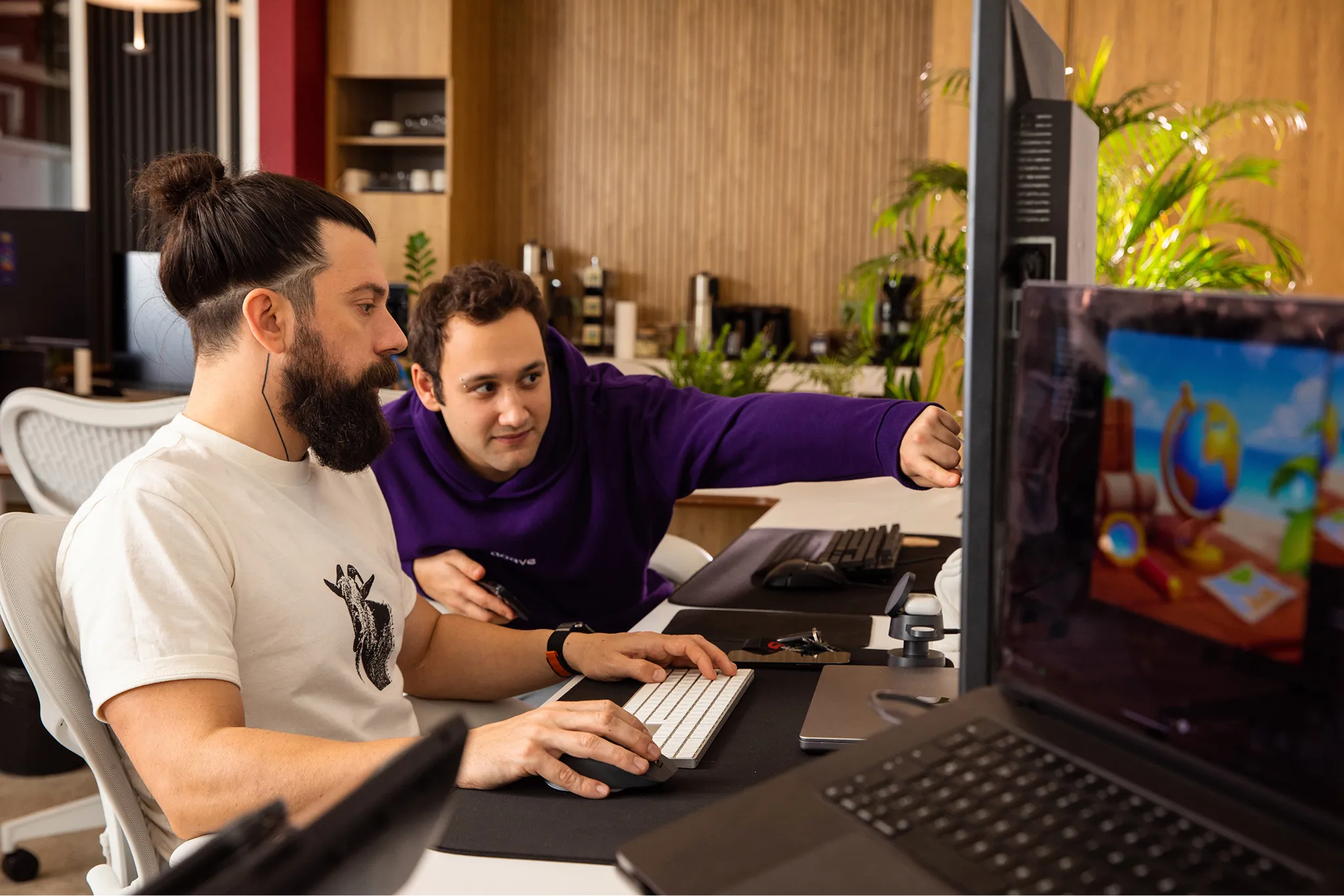 Two men working together at a desk in an office, one typing on a keyboard and the other pointing at a computer screen.
