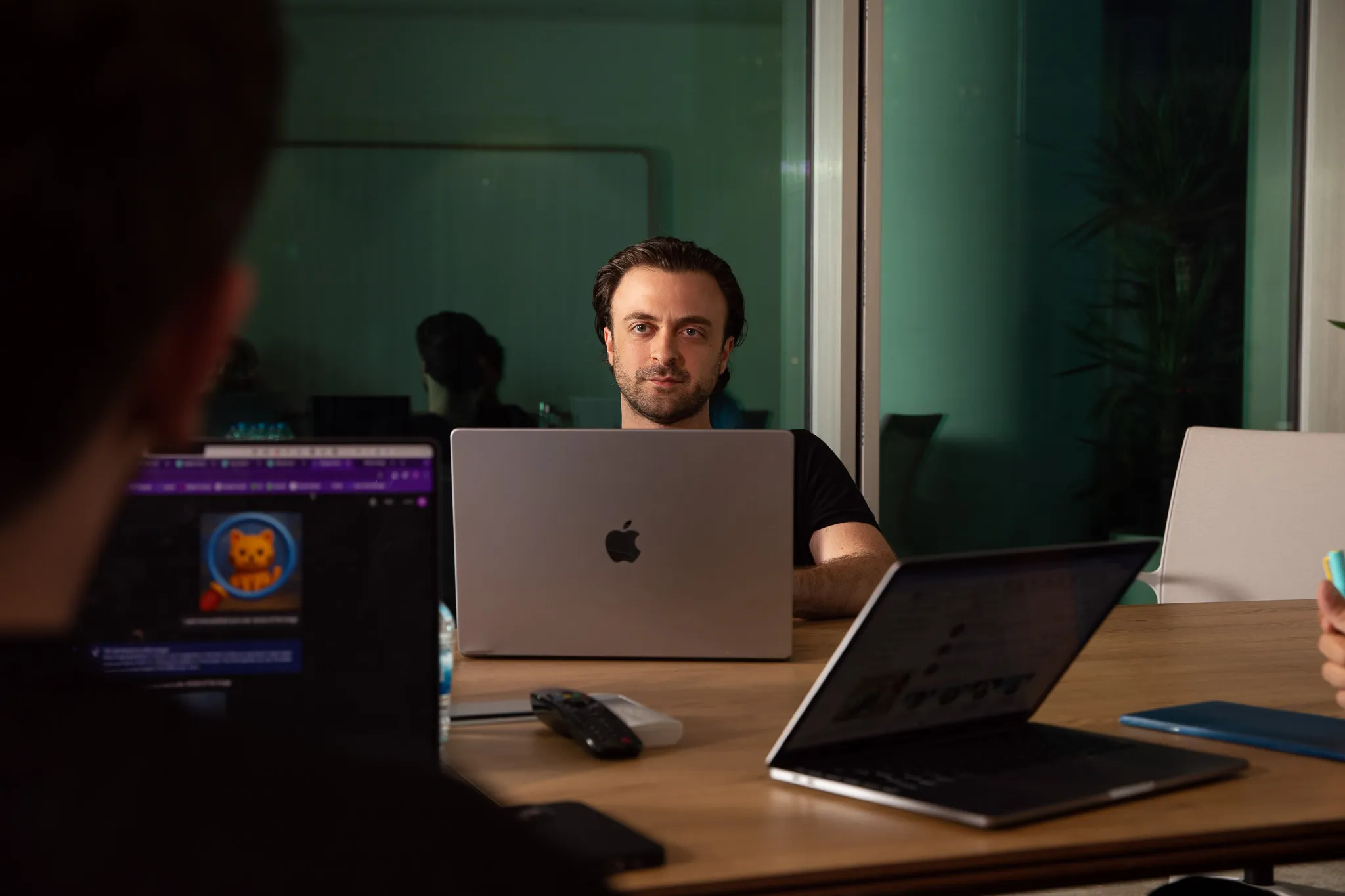 Man with dark hair sitting at a desk using a MacBook laptop, facing the camera during a meeting in an office.