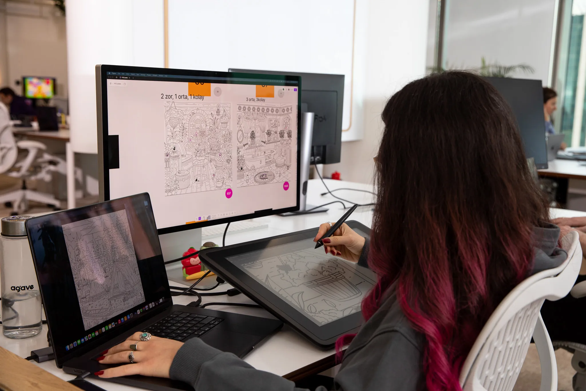 Woman with long dark hair dyed pink at the ends using a stylus to draw on a digital tablet beside a laptop and a monitor displaying black and white illustrations in an office setting.