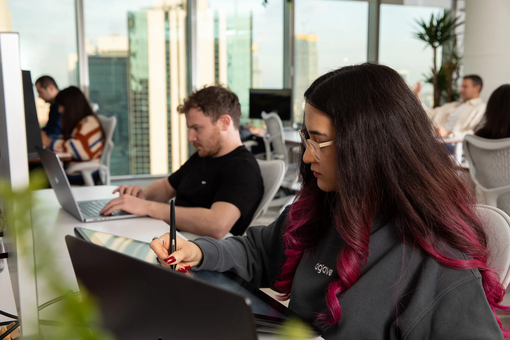 Woman with long dark hair and magenta highlights using a stylus on a digital drawing tablet in a modern office with colleagues working in the background.