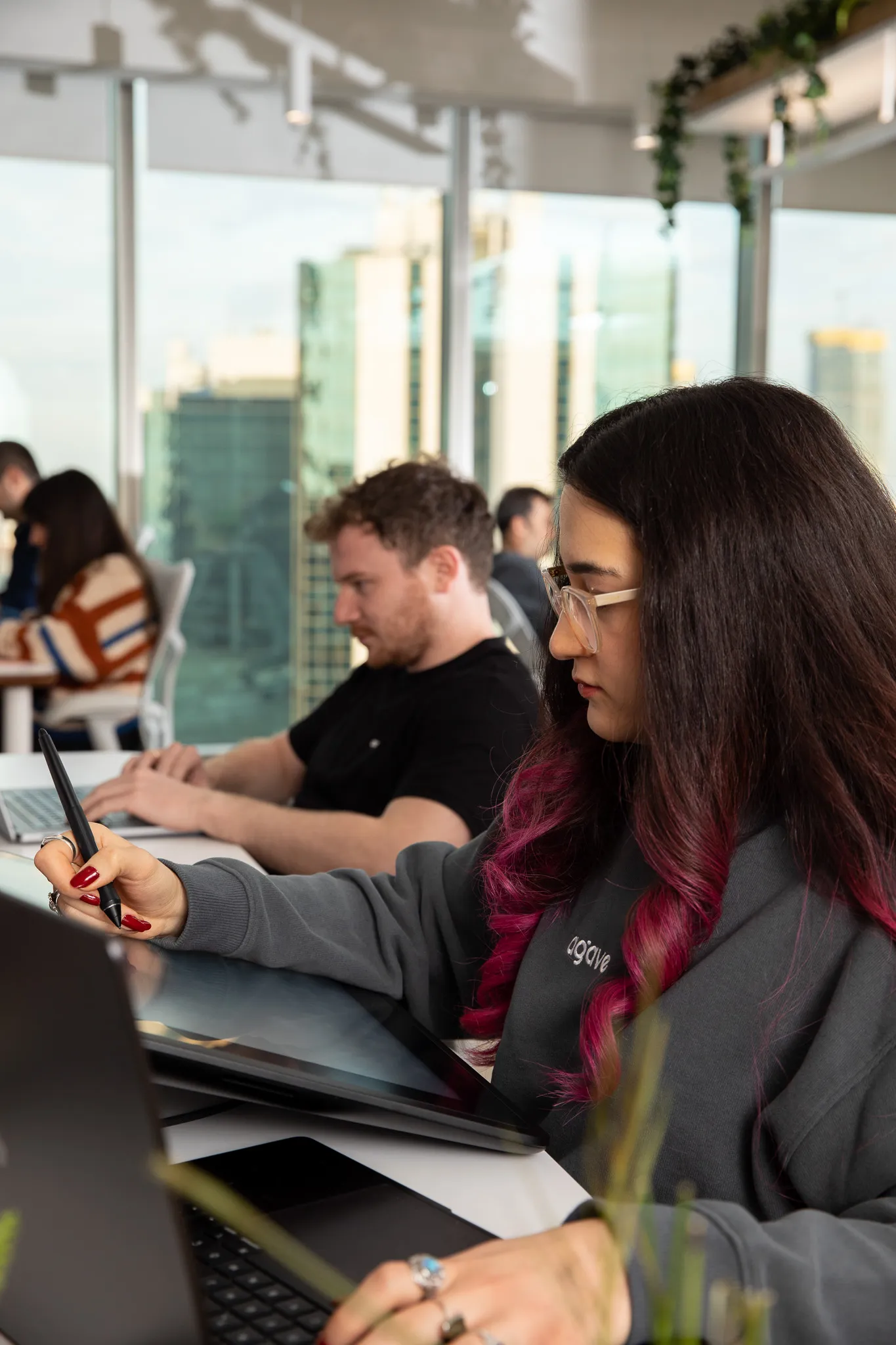 Woman with purple-tipped hair and glasses using a digital drawing tablet in a modern office with colleagues working on laptops in the background.