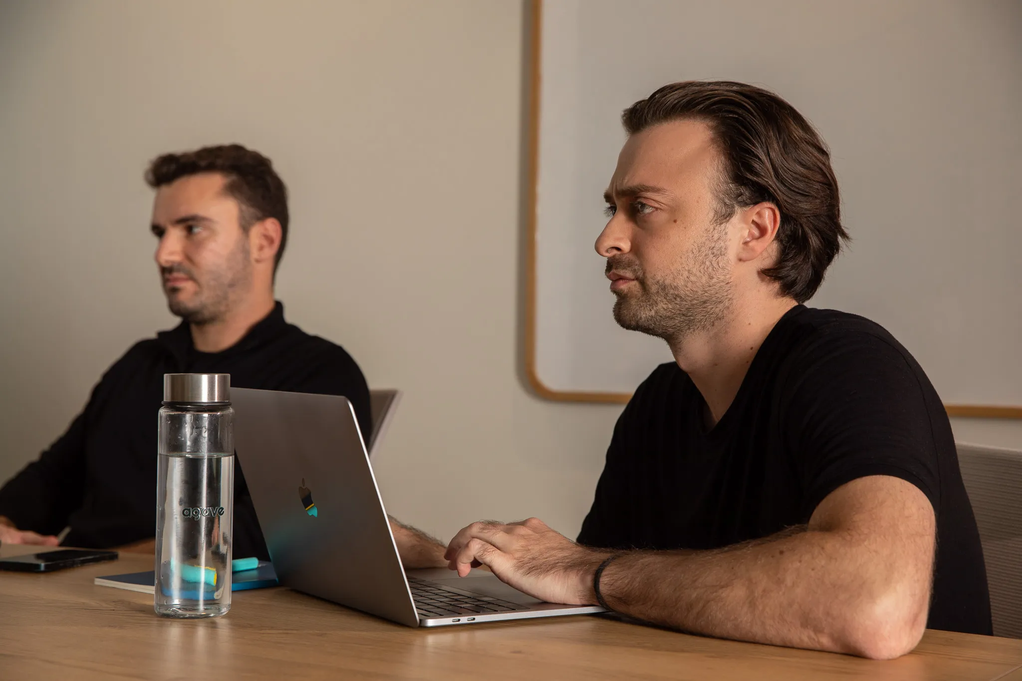 Two men sitting at a table in a meeting room, one typing on a laptop and the other looking ahead.