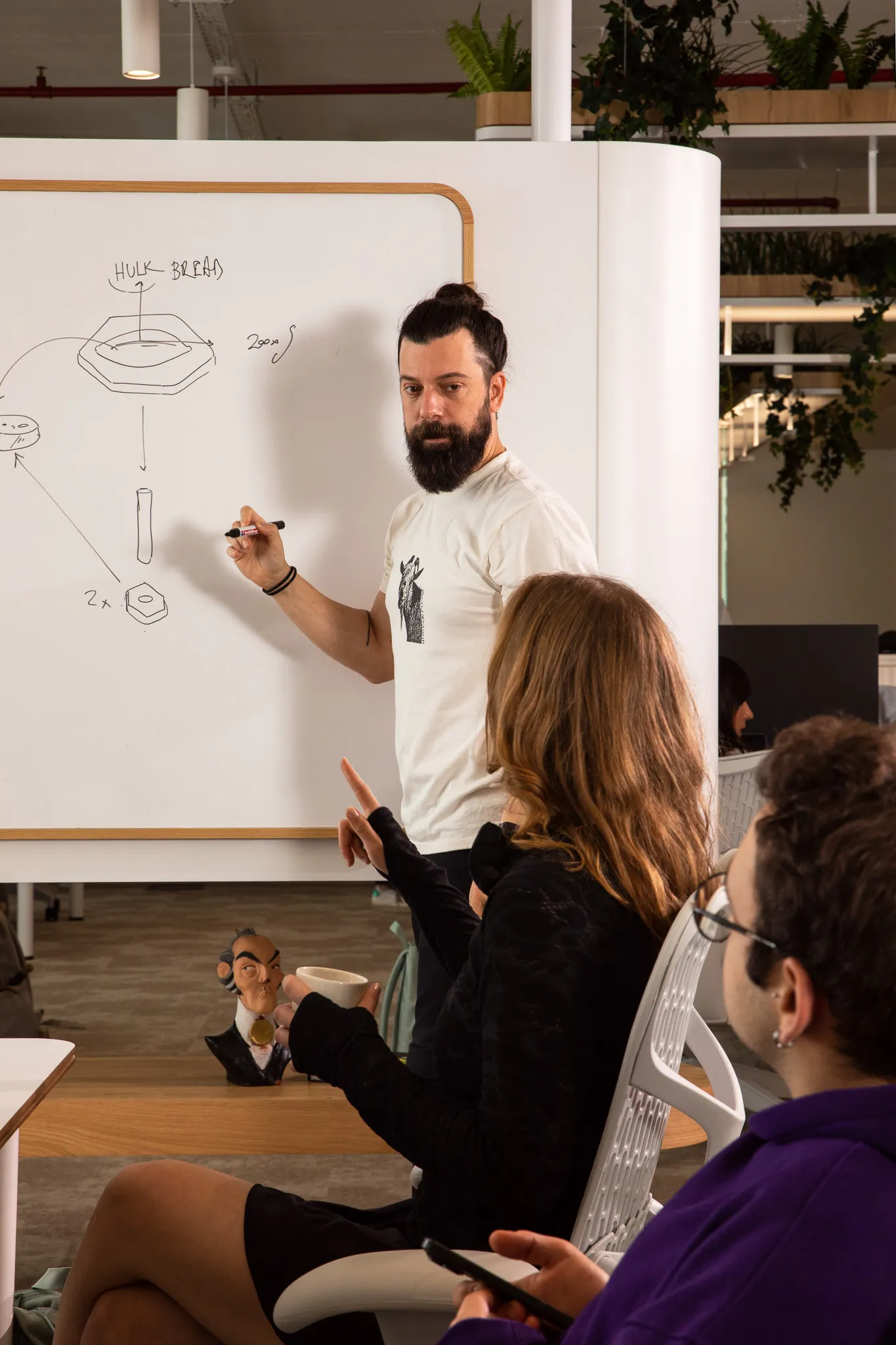 Bearded man with a bun explains a diagram on a whiteboard to two seated colleagues in an office.