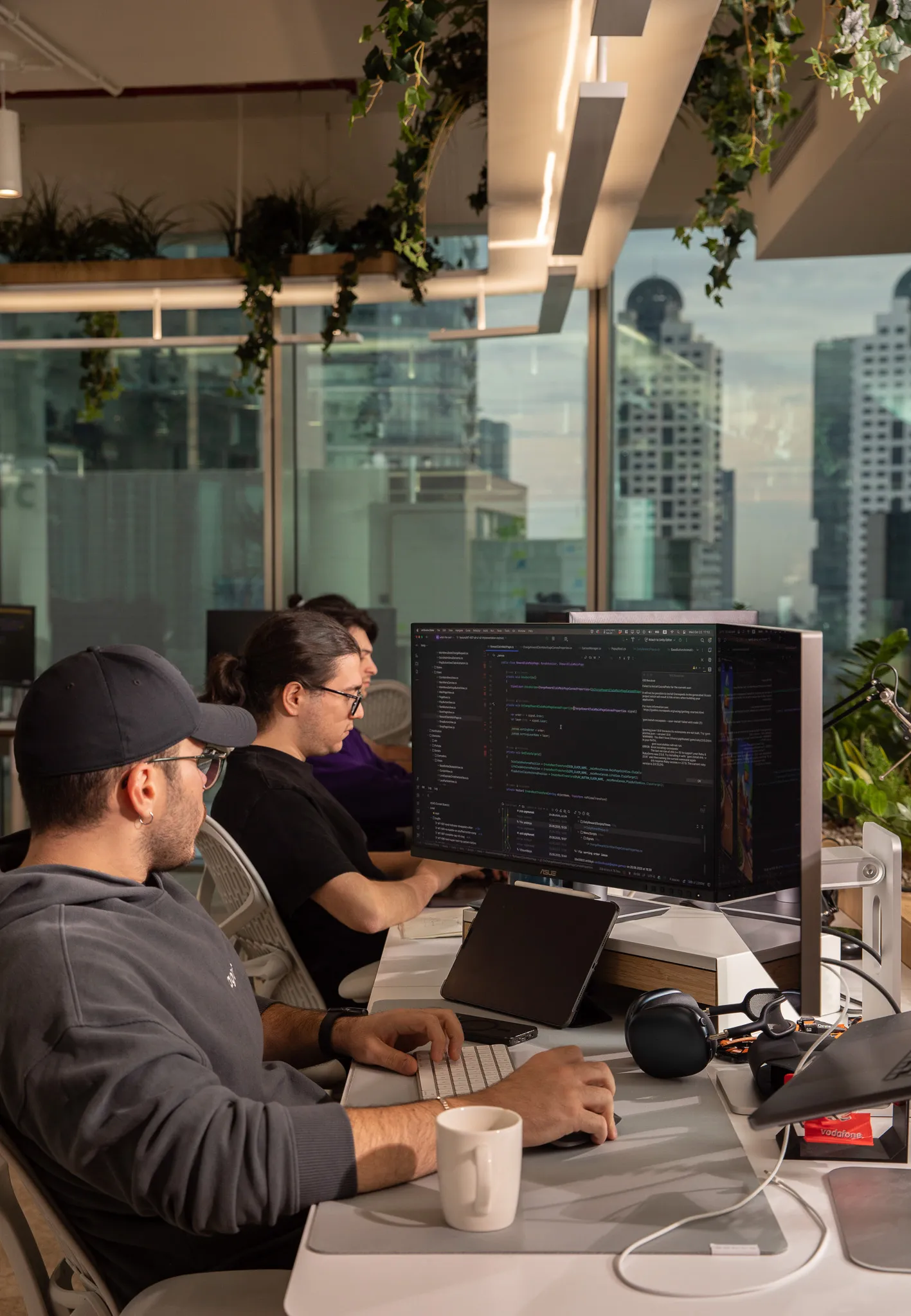 Three people working on computers at a modern office desk with a cityscape visible through large windows.