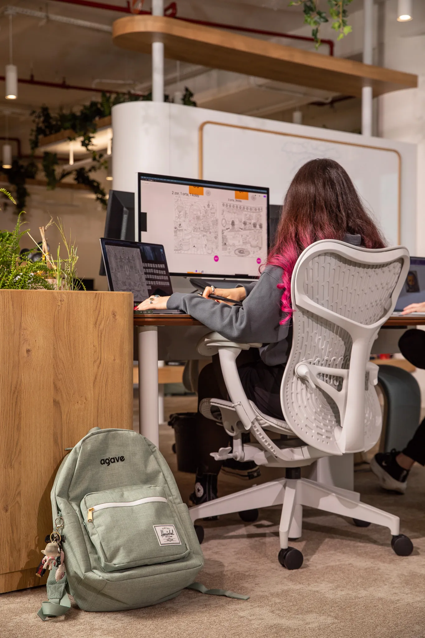 Person with long hair dyed pink at the ends working on dual monitors at a desk in a modern office, with a green backpack on the floor nearby.
