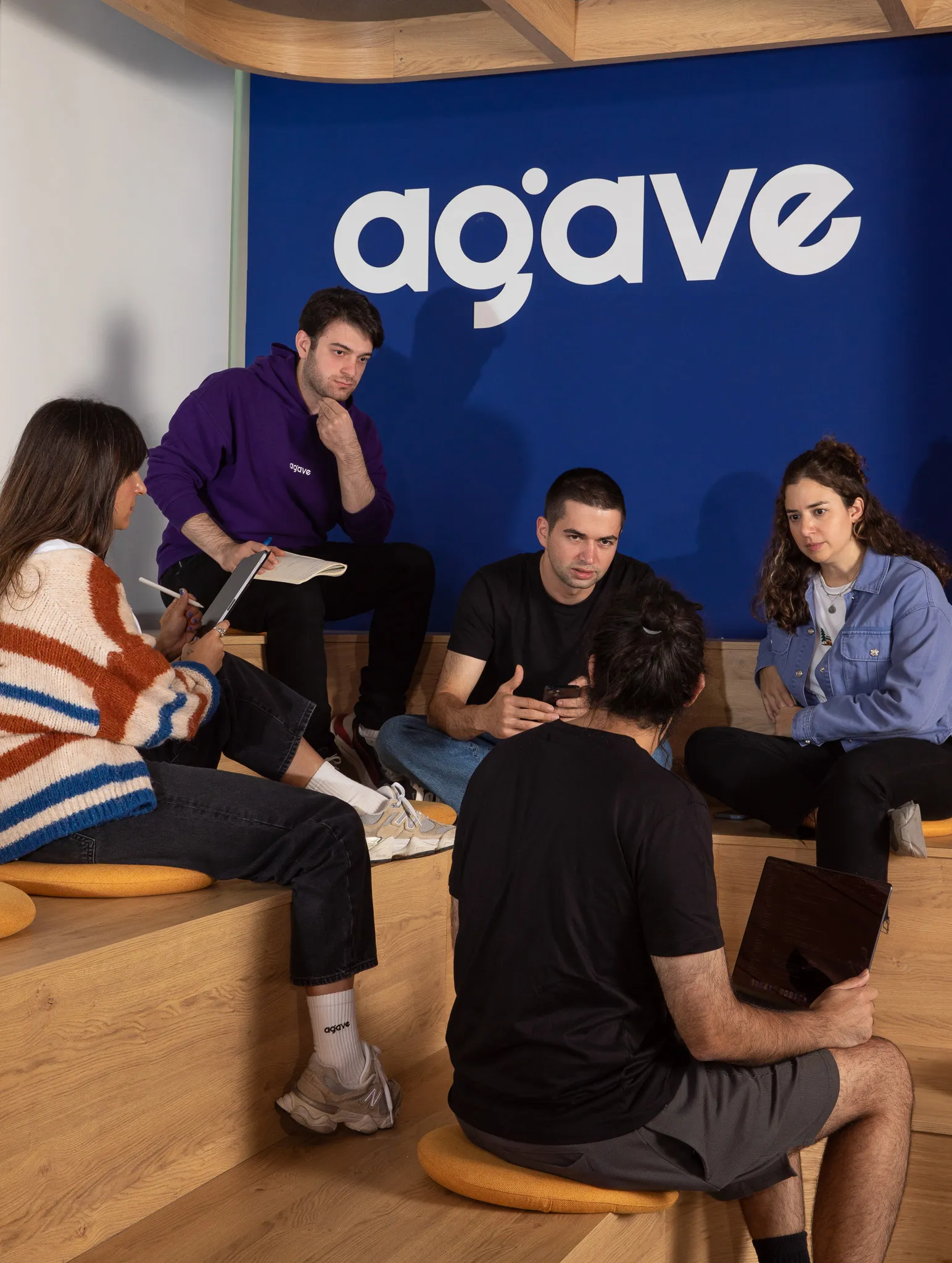 Five people engaged in a discussion seated on wooden stepped seating with a blue wall behind featuring the word 'agave'.