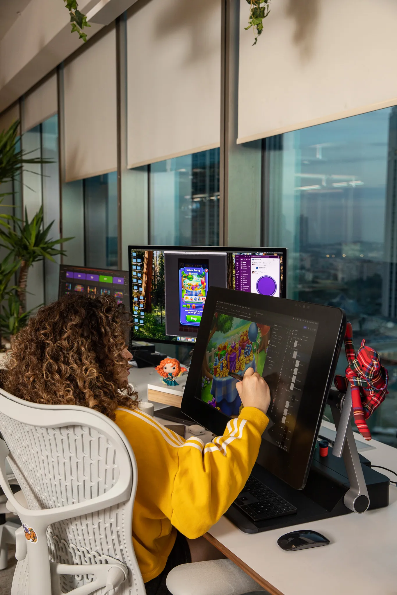 Person with curly hair in a yellow sweatshirt working on digital illustration of a colorful game on a pen display tablet at a desk with dual monitors in an office with large windows.