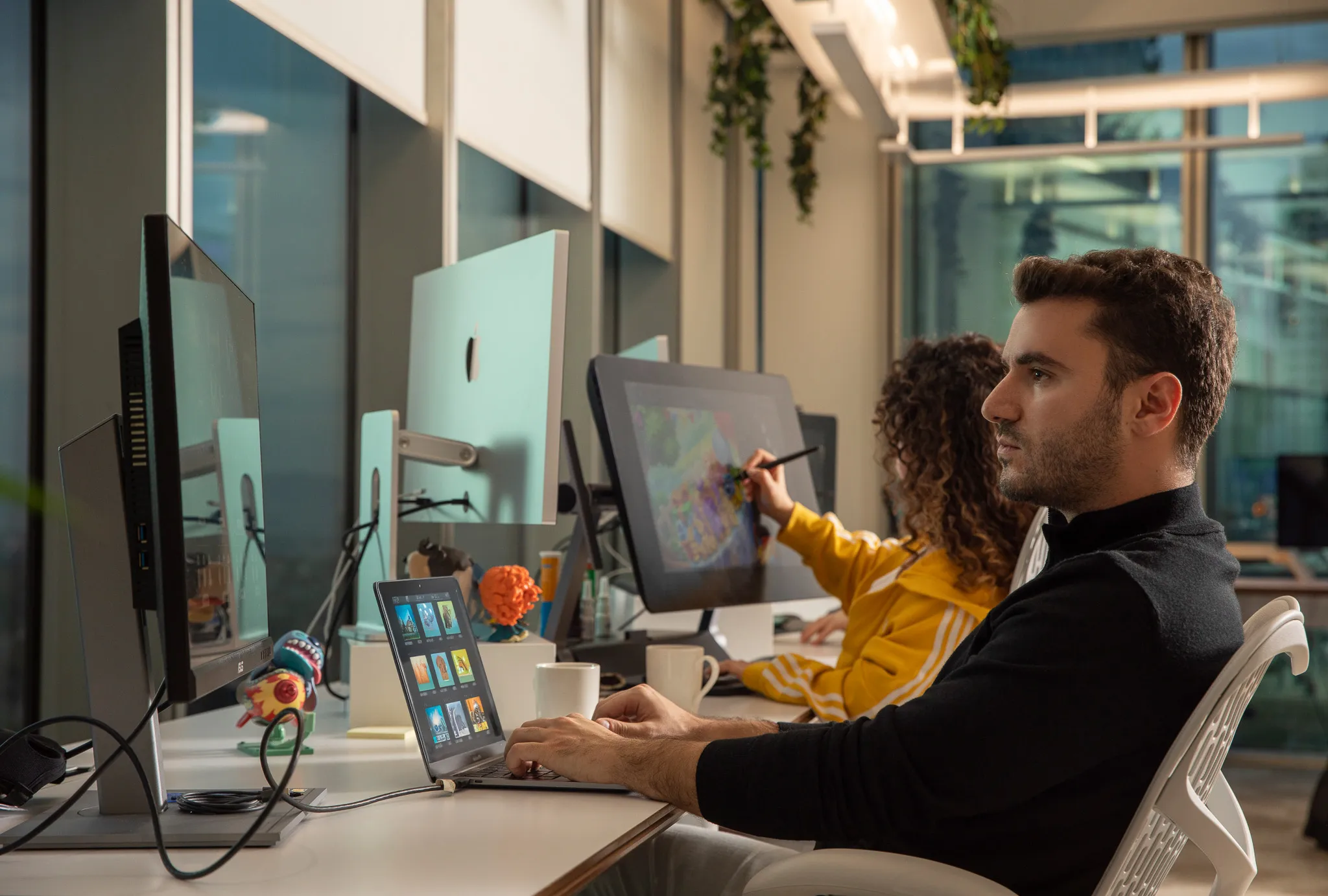Two people working at computers in an office, one using a laptop and the other drawing on a digital tablet.