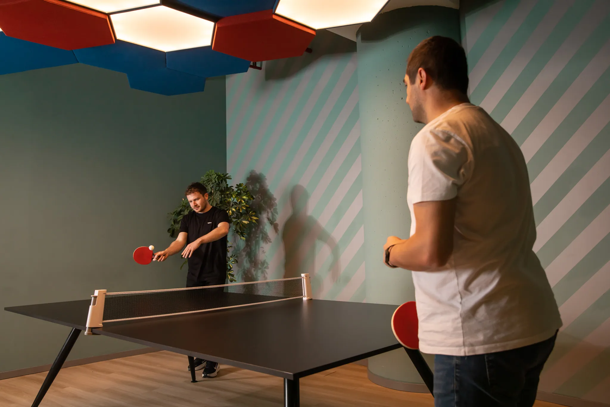 Two men playing table tennis indoors with a modern black table and red paddles.