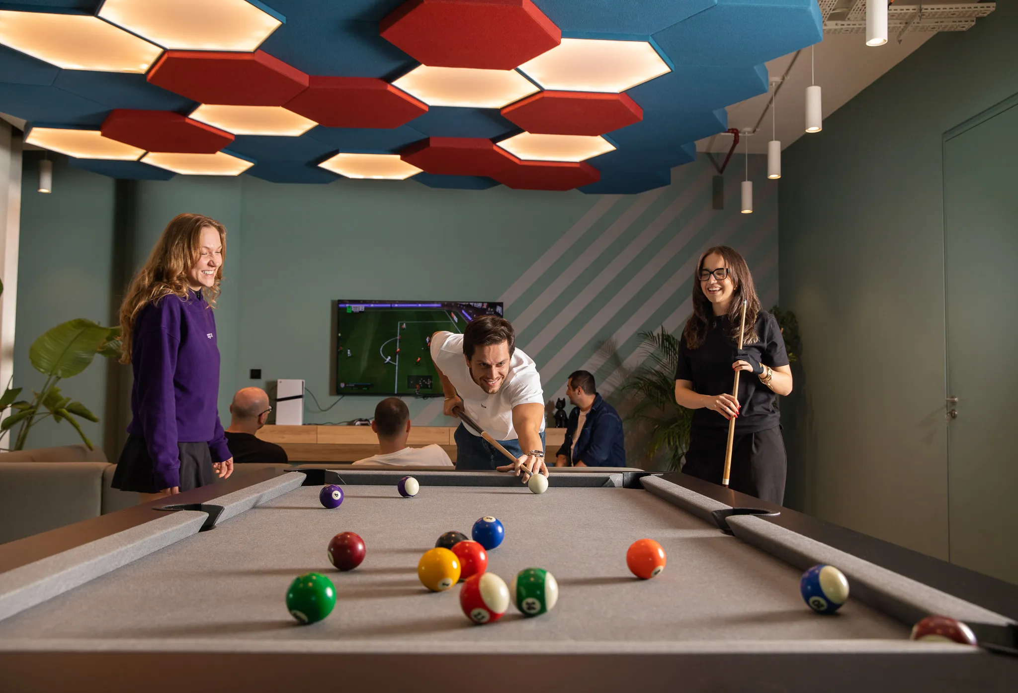 Three people playing pool on a table with colorful balls in a modern room with hexagonal ceiling lights and a TV in the background.