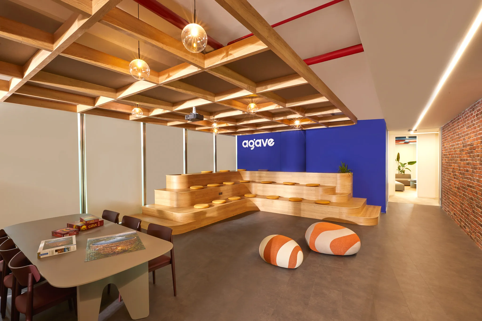 Modern office break area with wooden tiered seating, round orange and white cushions, a table with chairs, and a blue wall with 'agave' logo.