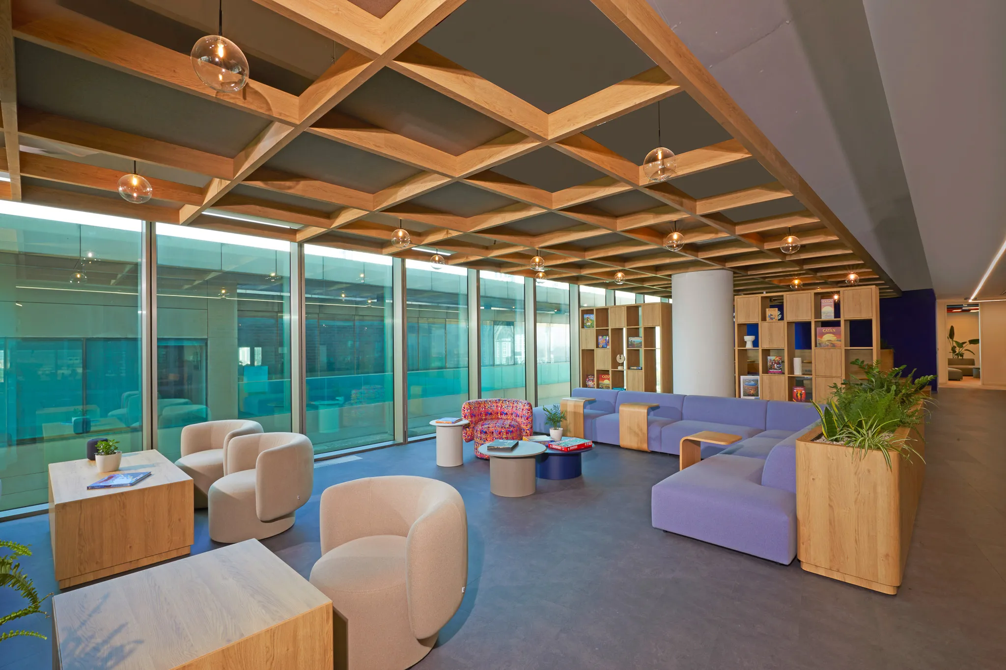 Modern lounge area with beige armchairs, purple sectional sofa, wooden tables, bookshelves, and large glass windows.
