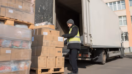 Warehouse worker in a safety vest loading cardboard boxes onto a pallet beside a large white delivery truck.