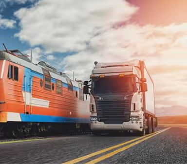 Orange-red train passing on railroad tracks beside a white semi-truck on a road under a cloudy sky with sunlight.