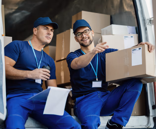 Two delivery workers in blue uniforms sitting in the back of a truck with boxes, one holding a clipboard and coffee, the other holding a cardboard box.