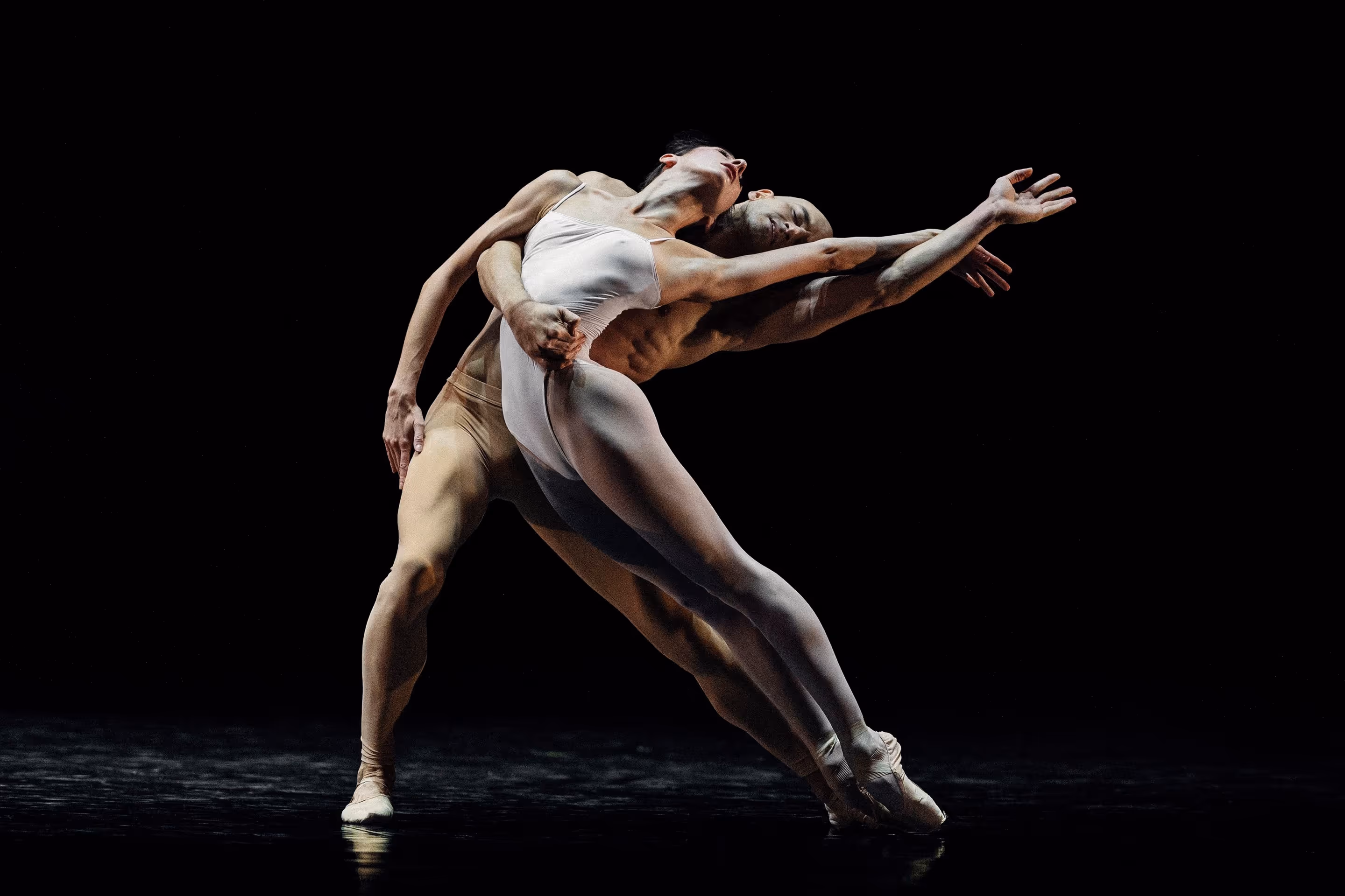 To balletdansere i kropstæt tøj udfører en elegant og dramatisk position mod en sort baggrund.