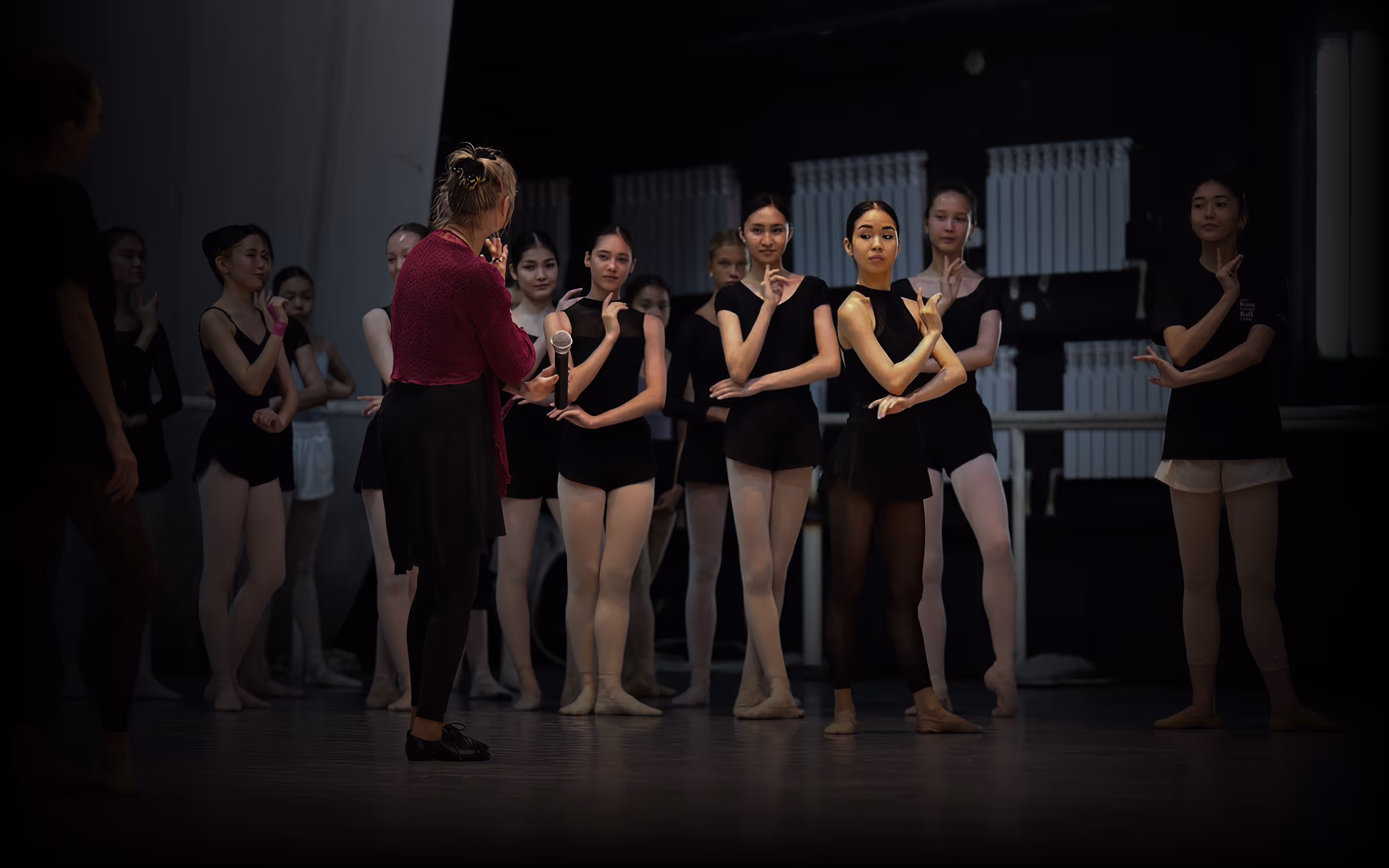 Balletinstruktør holder mikrofon og underviser en gruppe unge balletdansere i sorte dragt og strømpebukser under en dansetime.