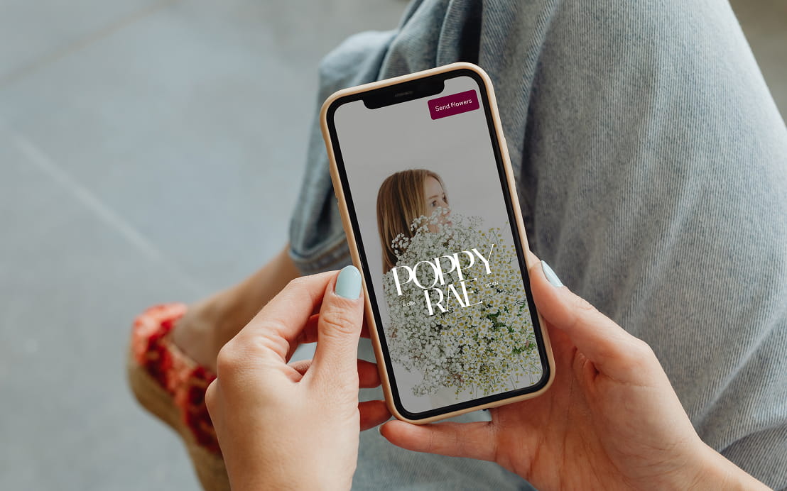 Person holding a smartphone displaying a floral app with a woman partially hidden behind a bouquet of white baby's breath flowers and the text 'POPPY RAE'.