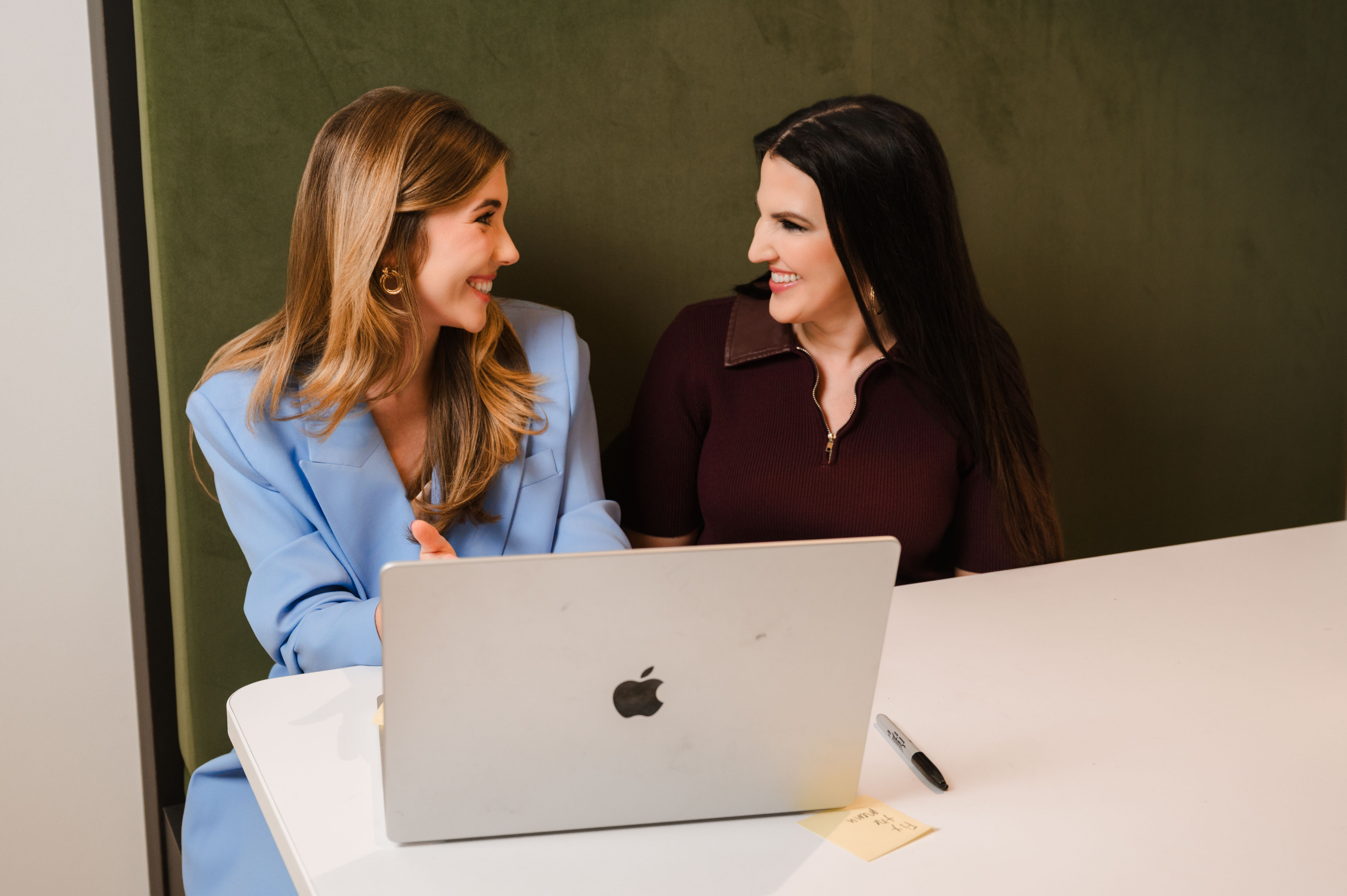Two women smiling and talking behind a laptop at a white table with a marker and sticky note.