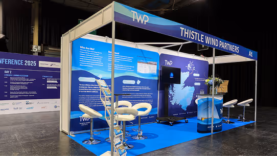 Exhibition booth for Thistle Wind Partners featuring informational panels, chairs, a TV screen, and a blue carpet at a conference venue.