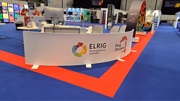 Exhibition booth with curved white panel displaying ELRIG and Drug Discovery logos on blue and red carpet.