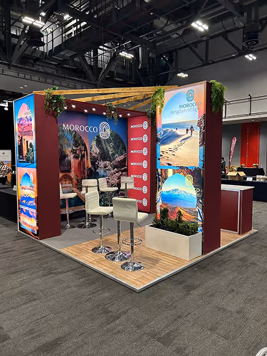 Exhibition booth showcasing Morocco tourism with colorful desert and mountain images, seating area, and greenery accents.