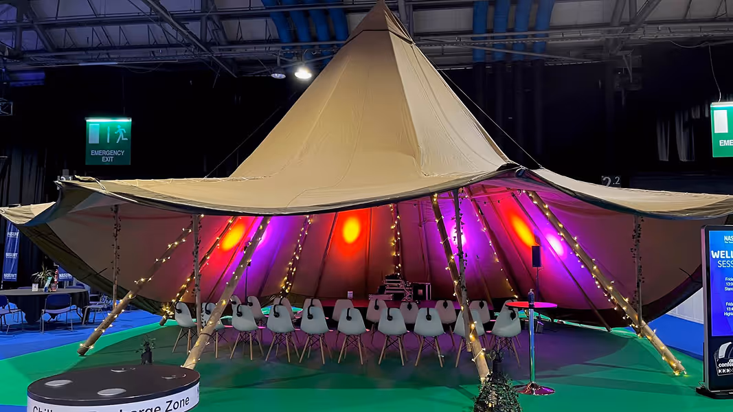 Large beige tent set up indoors with string lights wrapped around wooden poles and rows of white chairs inside, under colourful purple and orange lighting.