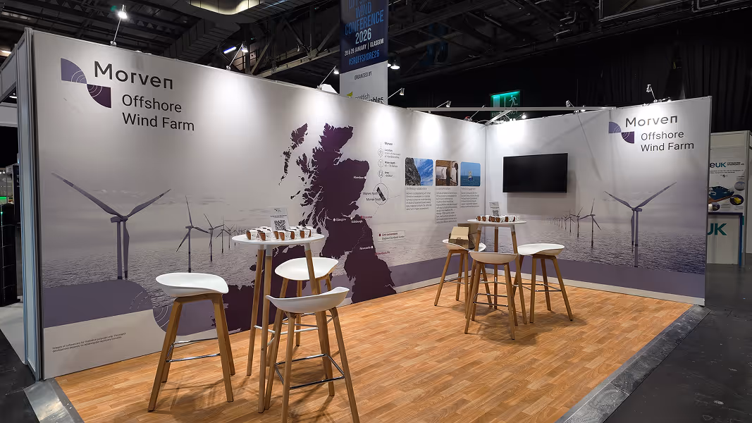 Exhibition booth for Morven Offshore Wind Farm featuring a map of the United Kingdom, informational panels, and tall tables with white stools on a wood-patterned floor.