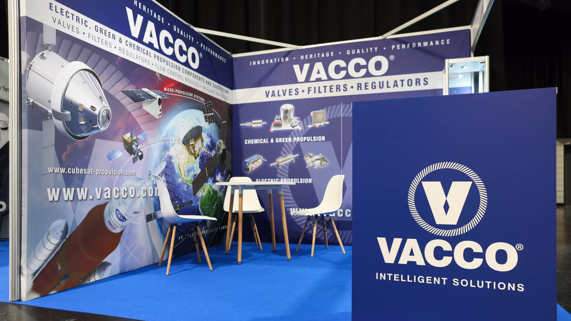 Exhibition booth for VACCO featuring space propulsion graphics, blue floor, table, and white chairs.