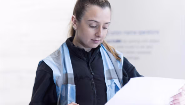 Woman in a work vest reading a document with focus indoors.