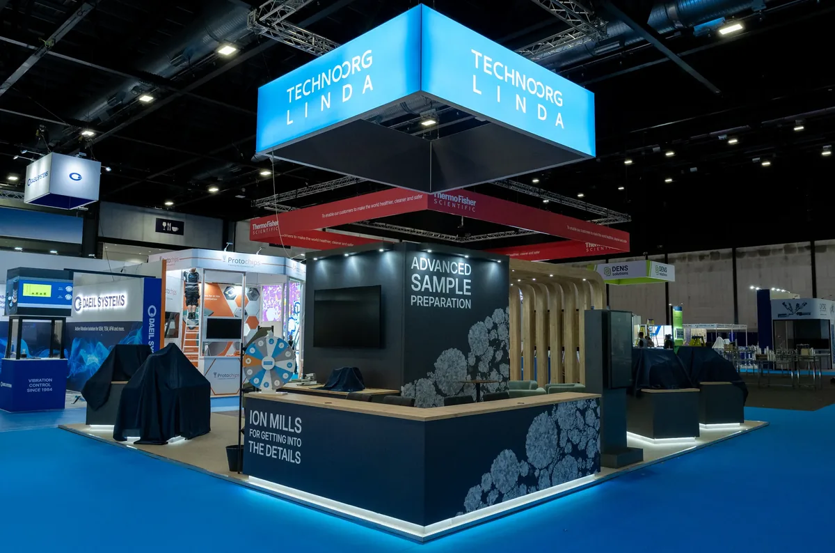 Trade show booth for Technoorg Linda featuring illuminated blue hanging signs with the company name, black display walls with text about advanced sample preparation and ion mills, and a modern setup with covered equipment and seating areas.
