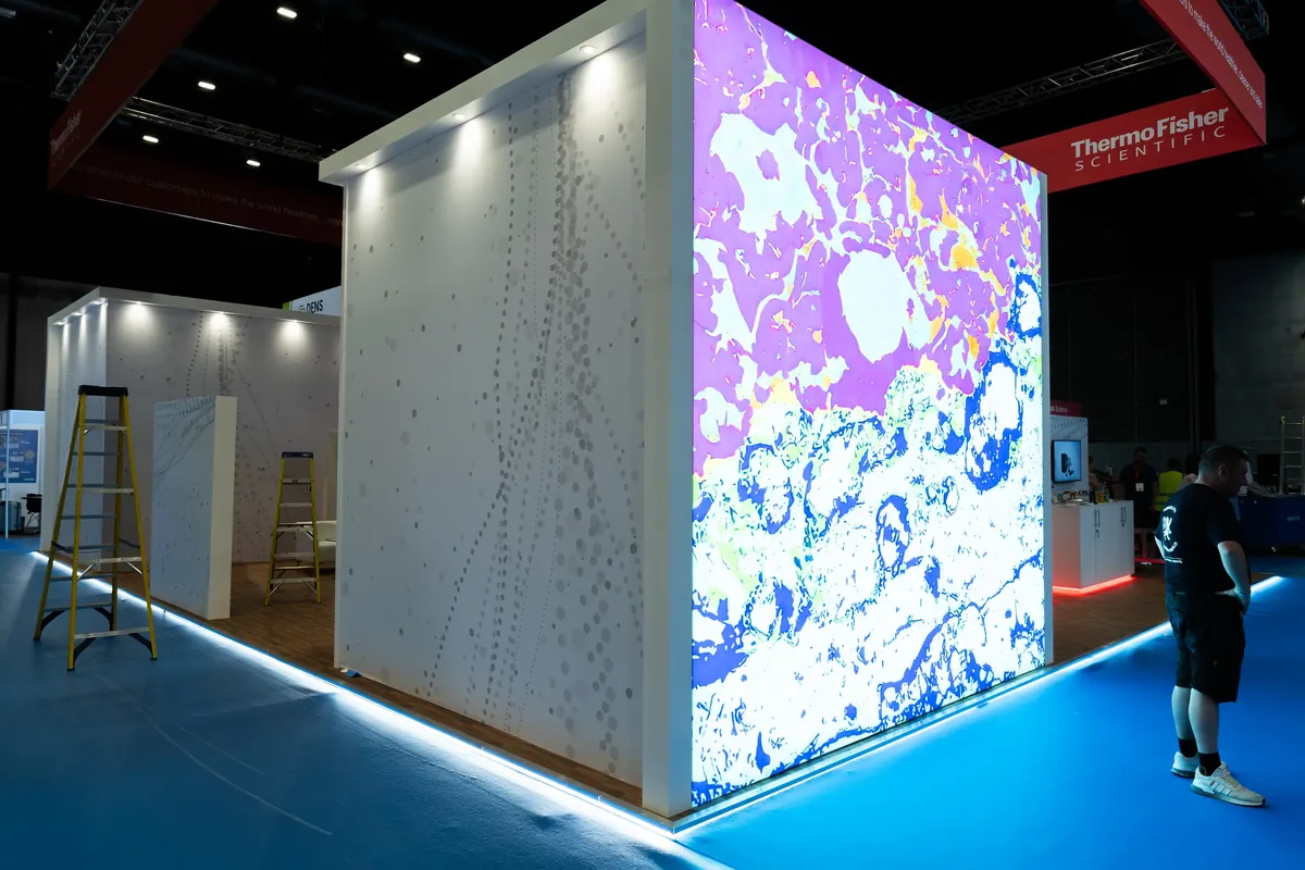 Exhibition booth with large illuminated screen showing colorful scientific imagery in pink, white, and blue; two yellow ladders and a person in casual clothes standing on bright blue floor; Thermo Fisher Scientific banner overhead.